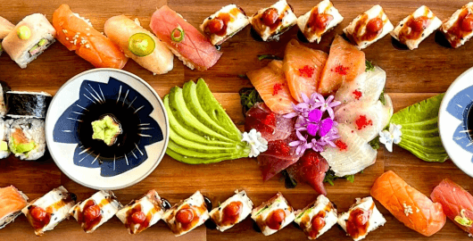 A large wooden catering board covered with a variety of fresh sushi rolls, nigiri, sashimi arrange with edible flowers, sliced avocado, and soy sauce.