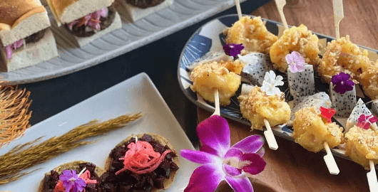 An elegant catering platter with glazed skewers garnished with edible flowers and dragon fruit, alongside sliders and small bites on a wooden table.